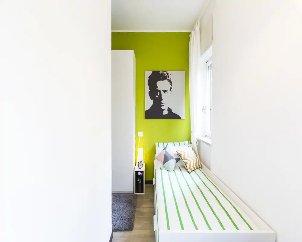 Chambre simple lumineuse avec un lit blanc aux draps rayés verts, coussins décoratifs, lampe de chevet et une œuvre moderne sur un mur vert vif. Pièce compacte, propre et bien éclairée par la lumière naturelle.