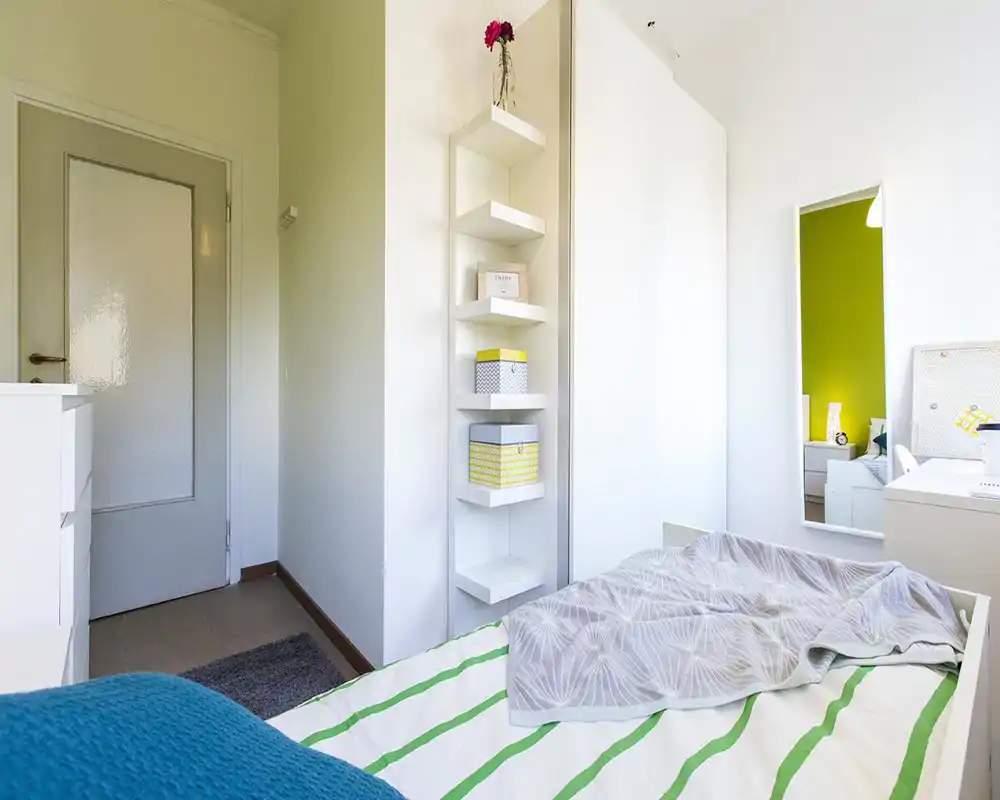 Chambre lumineuse et minimaliste avec un lit simple bien fait, des meubles blancs et des étagères décoratives ; une grande glace reflète la pièce et une paroi vert citron est visible en reflet.