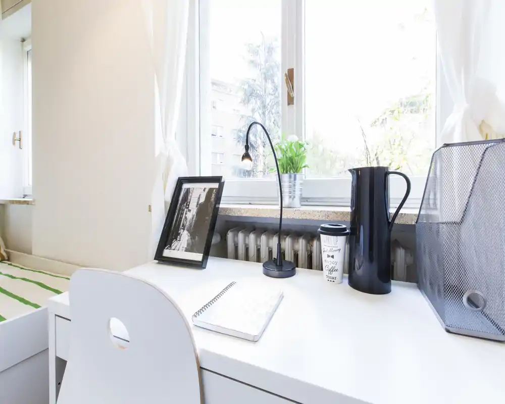 Chambre lumineuse et minimaliste avec un coin bureau devant une grande fenêtre : bureau blanc, chaise et quelques éléments décoratifs comme une plante et une lampe.