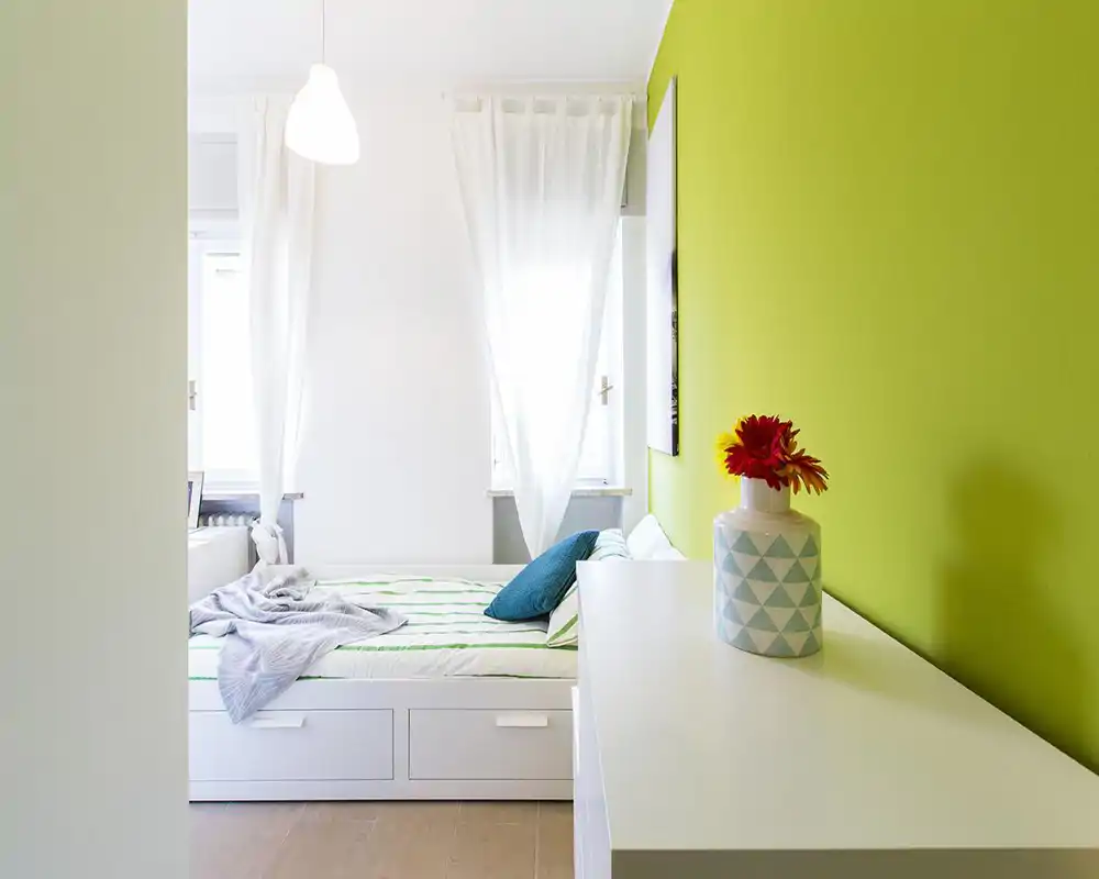 Chambre moderne et lumineuse avec lit de rangement blanc, linge doux et mur d'accent vert. La lumière naturelle de deux fenêtres et les touches décoratives rendent l'espace frais et accueillant.