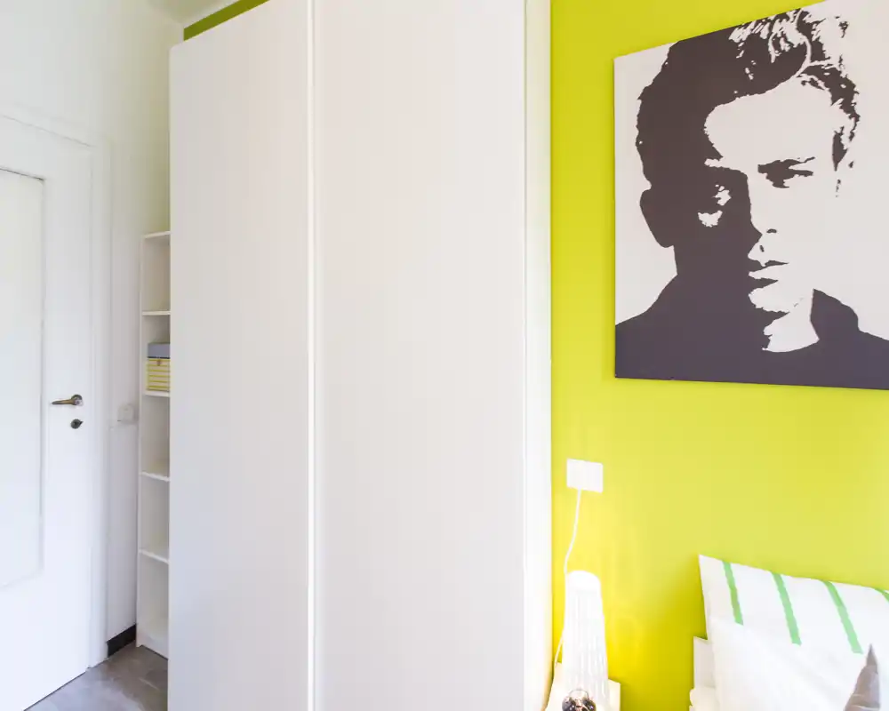 Coin de chambre lumineux et moderne avec une armoire blanche, une lampe de chevet et un mur vert ; seul un coin du lit et des oreillers est visible.