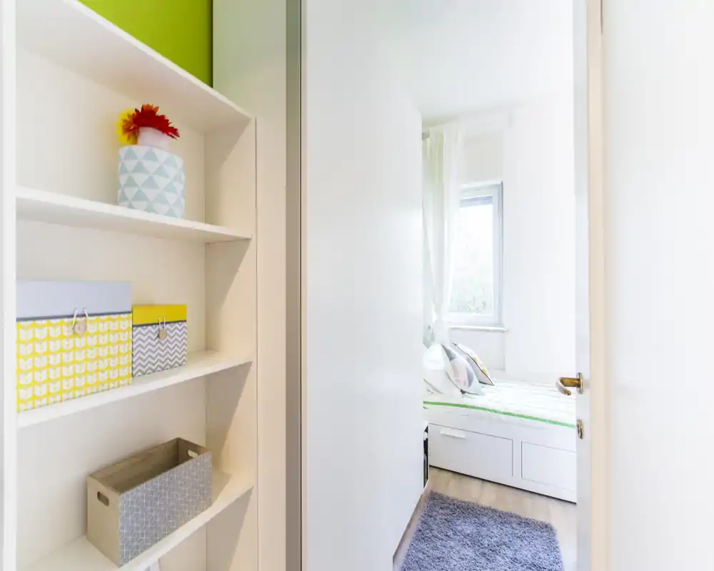 Chambre lumineuse et compacte vue par une porte ouverte, lit simple avec coussins et tapis moelleux ; style moderne et épuré, parfait pour une petite location.