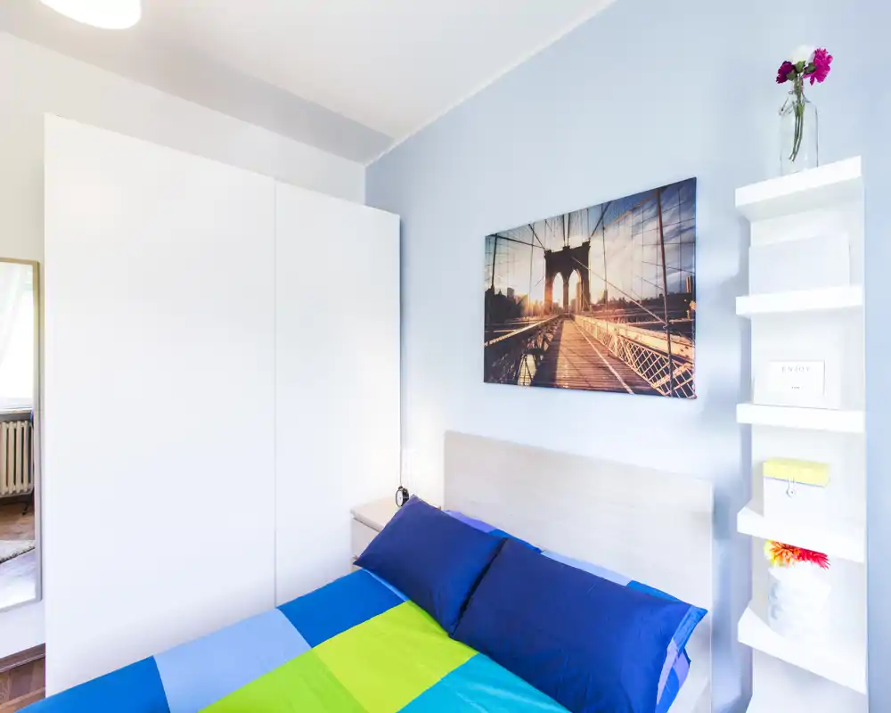 Camera da letto luminosa e ben arredata con un letto matrimoniale con biancheria colorata, un ampio armadio bianco e quadri a parete: perfetta per annunci in affitto.