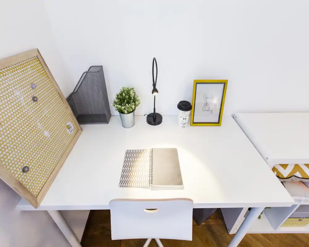 Angolo studio luminoso e minimale con scrivania bianca, lampada, quaderno e pianta decorativa—perfetto per studio o lavoro da remoto.