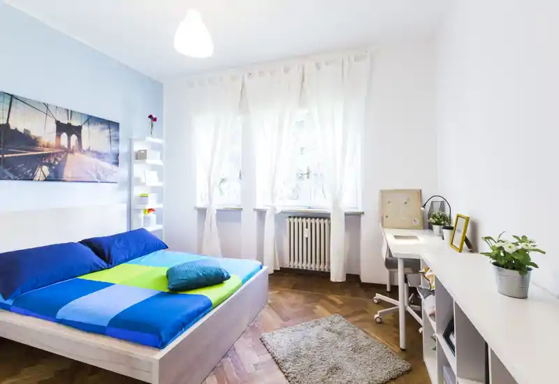 Camera da letto moderna e luminosa con letto matrimoniale, biancheria colorata, luce naturale da una finestra ampia e una scrivania ordinata — perfetta per un soggiorno confortevole.