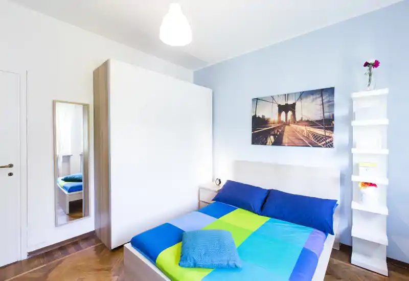 Camera da letto moderna e luminosa con un letto matrimoniale ben rifatto, biancheria colorata e un ampio armadio. Parete azzurra, stampa decorativa sopra la testata e specchio a figura intera.