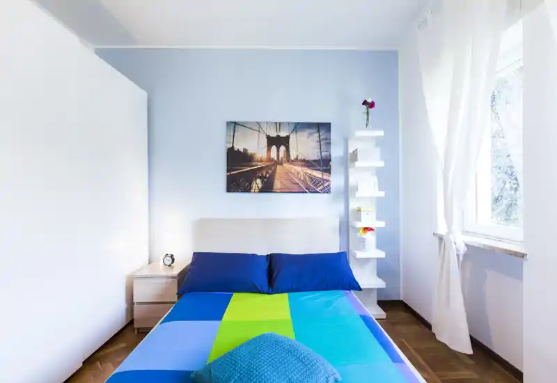 Camera da letto luminosa e moderna con letto matrimoniale con biancheria colorata, testiera minimal, quadro a parete e finestra che illumina bene l'ambiente.