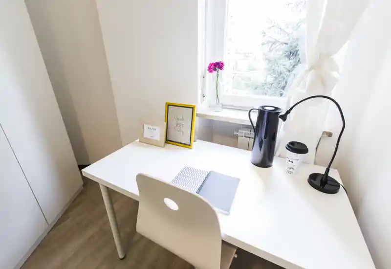 Bright, minimal desk corner with a white table, chair, desk lamp and small decorative items next to a window — ideal for a quiet workspace within the living area.