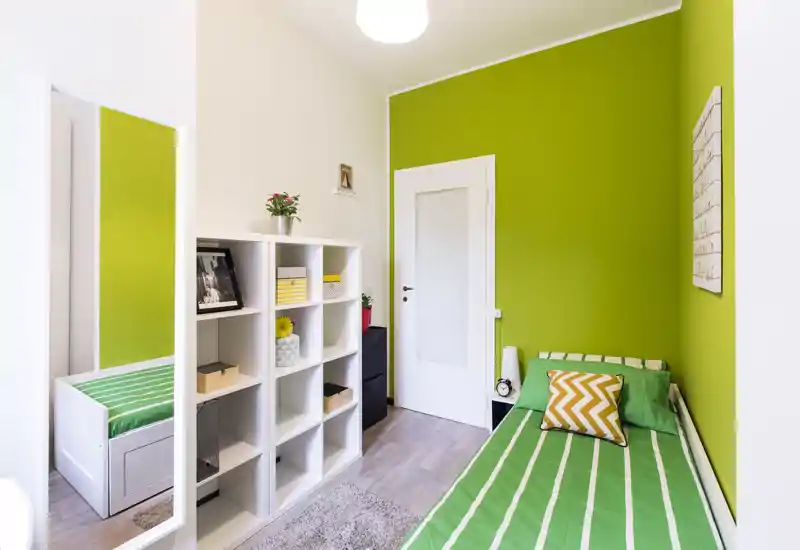 Bright, neatly staged single bedroom with a green accent wall, striped bed linen, shelving unit and decorative pillow — ideal for a student or short-term rental.