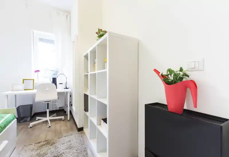 Compact, bright bedroom with a small desk by the window, open shelving and a partially visible bed. Neutral tones, a soft rug and plants create a fresh, functional feel ideal for a student or single occupant.