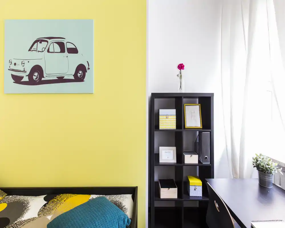 Angolo di camera luminoso e moderno con letto parzialmente visibile, plaid colorato e stampa grafica; una libreria e una scrivania creano un'area lavoro ordinata vicino alla finestra.