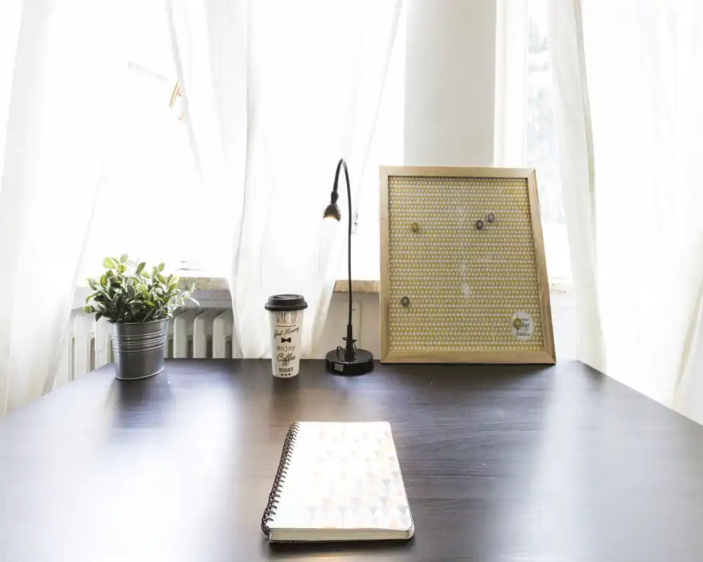Vista di un angolo living luminoso e minimale con un tavolo/desk ordinato vicino alla finestra: pianta, lampada e pannello decorativo rendono l’ambiente accogliente per un’area lavoro in soggiorno.