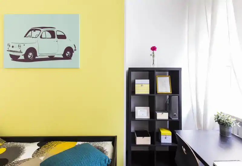 Angolo di camera luminoso e moderno con letto parzialmente visibile, plaid colorato e stampa grafica; una libreria e una scrivania creano un'area lavoro ordinata vicino alla finestra.