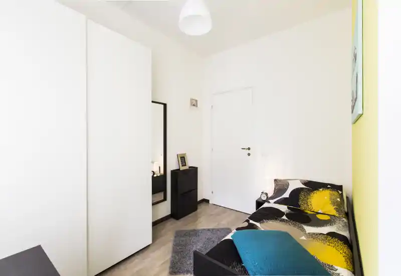 Camera da letto singola luminosa e moderna con letto dai motivi grafici, cuscino turchese e armadio bianco minimal — compatta e accogliente.