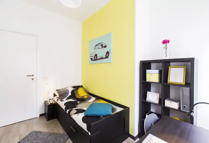 Camera luminosa e compatta con letto singolo con struttura nera, cuscini colorati e una moderna libreria a cubi — ideale per studenti o affitti brevi.