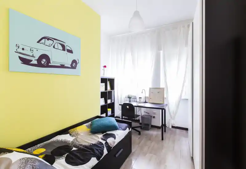 Camera da letto luminosa e arredata con un letto singolo, parete colorata e una scrivania vicino alla finestra — ideale per uno studente o una persona sola.