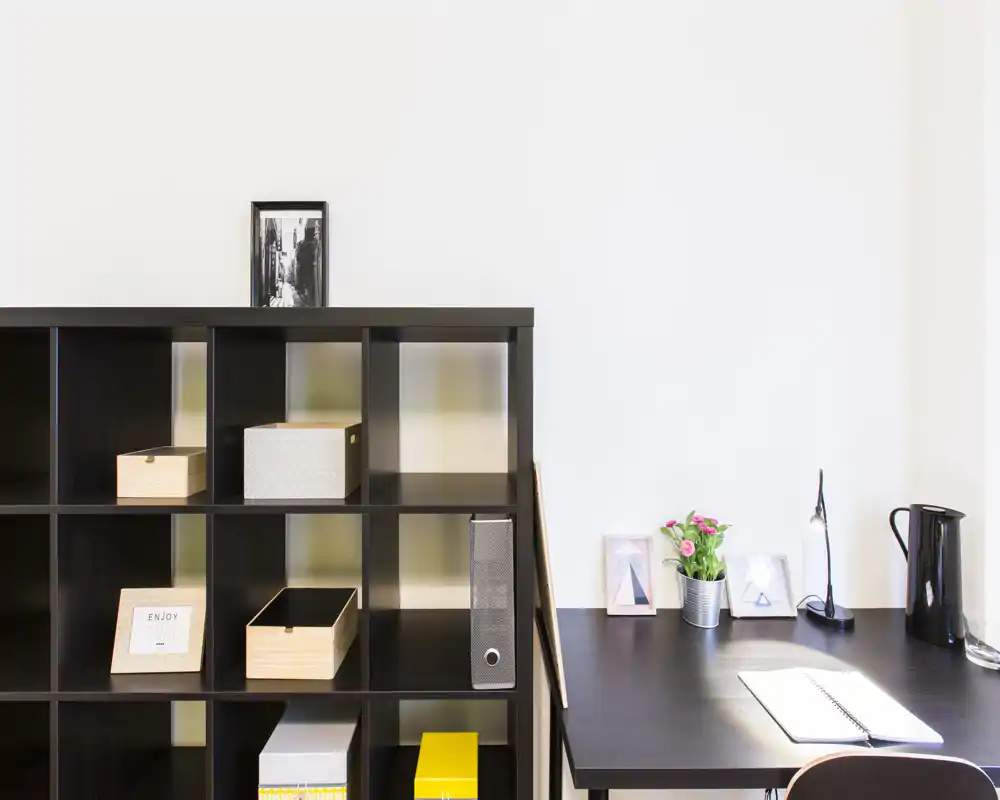 Rincón de salón moderno y ordenado con estantería oscura y un escritorio limpio. Cajas decorativas, fotos enmarcadas y una pequeña planta crean un ambiente minimalista y acogedor para trabajar o relajarse.