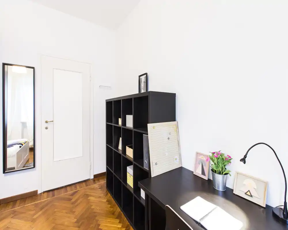 Habitación luminosa de uso múltiple con estantería oscura y escritorio, decorada con cuadros, una planta y una lámpara — ideal como oficina en casa o zona de estar.