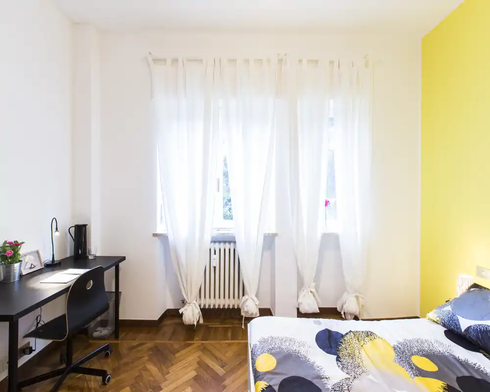 Dormitorio luminoso y moderno con ropa de cama con estampado, escritorio y ventana soleada. La pared amarilla y el parquet aportan calidez y carácter.