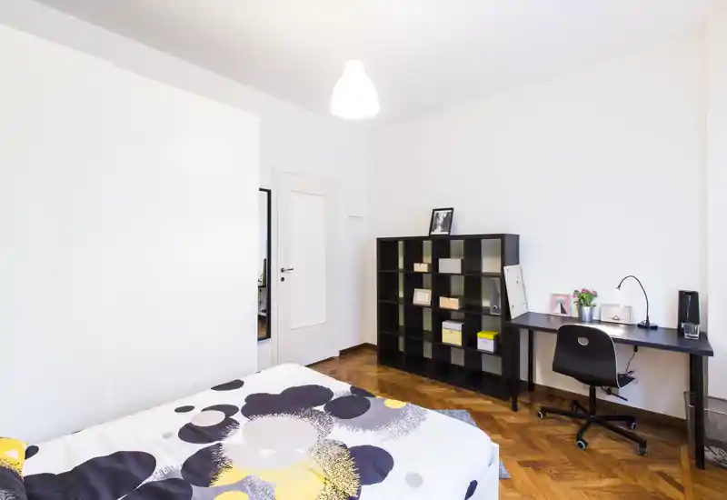 Dormitorio moderno y luminoso con edredón estampado, suelo de parquet, estantería y una mesa de trabajo compacta — perfecto para una estancia cómoda o teletrabajo.