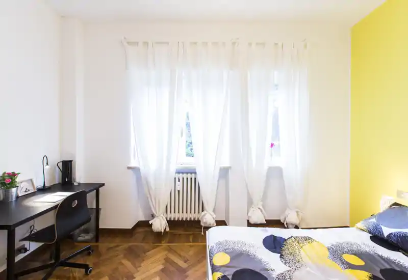 Dormitorio luminoso y moderno con ropa de cama con estampado, escritorio y ventana soleada. La pared amarilla y el parquet aportan calidez y carácter.