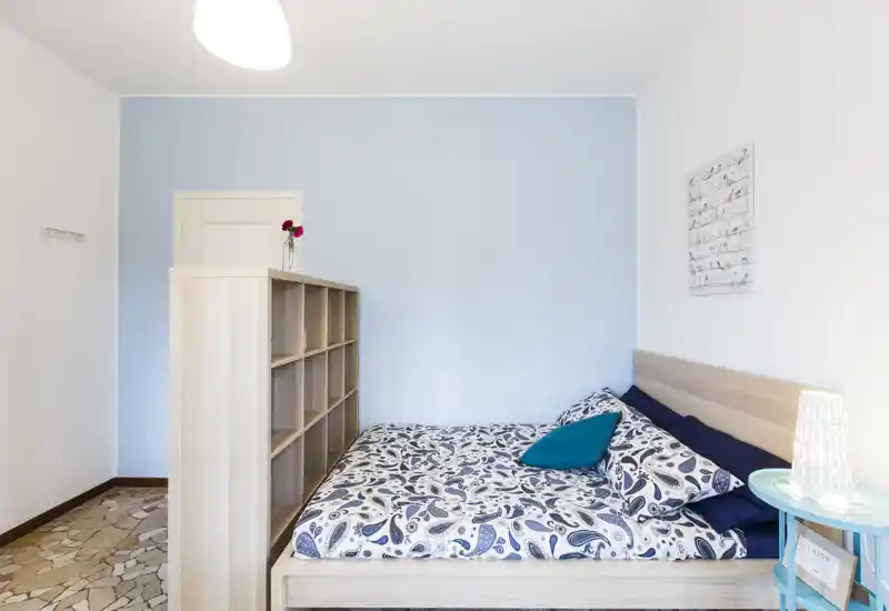 Camera da letto luminosa e ben arredata con biancheria stampata, testiera in legno e libreria divisoria, ideale per un soggiorno accogliente in singola o doppia.