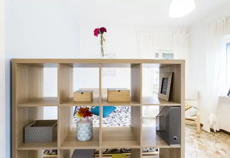 Camera da letto luminosa e moderna con una scaffalatura decorativa in primo piano e un letto con fodere a motivo e cuscini visibile dietro.
