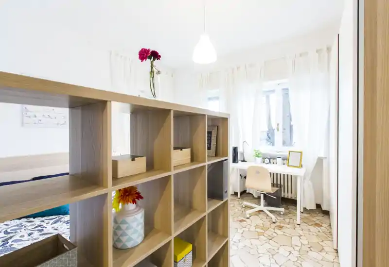 Camera da letto moderna e luminosa con una libreria divisoria in legno, zona letto visibile e un piccolo angolo studio vicino alla finestra. L'ambiente risulta arioso grazie alle tende bianche e alla luce naturale.
