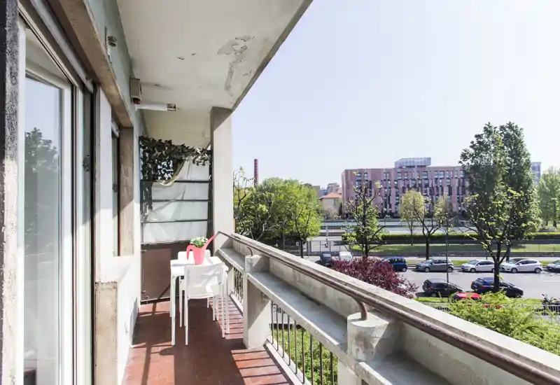 Sunny balcony with a small table and two chairs, overlooking a tree-lined street and modern buildings — ideal for morning coffee.