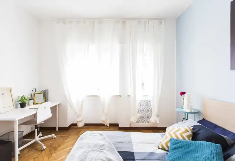Bright, modern bedroom with a neatly made bed, soft natural light from the large window, and a compact desk — ideal for a comfortable stay or remote work.