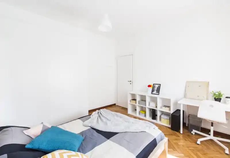 Bright, minimalist bedroom with a large bed, colorful cushions and a small desk area by the window. Clean parquet flooring and white walls create a fresh, airy atmosphere ideal for rental photos.