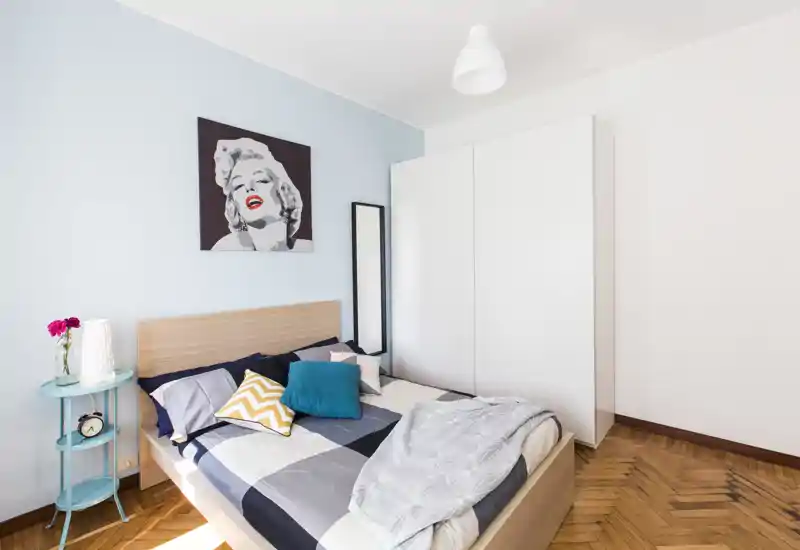 Bright, modern bedroom with a double bed, colorful cushions and a white wardrobe; tasteful artwork and parquet flooring enhance the space.