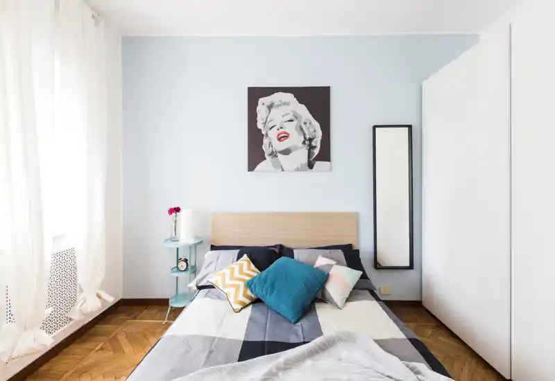 Bright, stylish bedroom with a double bed, colorful cushions and a portrait artwork above the headboard. Large window with light curtains and parquet flooring create a warm, airy feel ideal for rental listings.