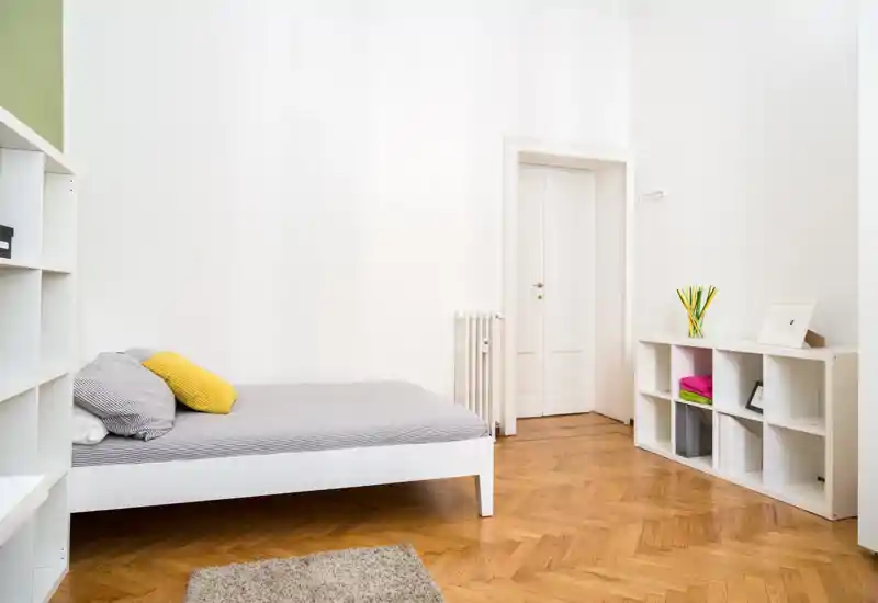 Minimalist bedroom with a low white bed, gray bedding and a yellow accent cushion; bright room with parquet flooring and simple shelving for storage.