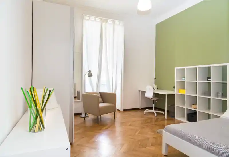 Bright, modern bedroom with a neatly made bed, a cosy armchair by the window, a study desk and open shelving — ideal for single occupancy or a student.