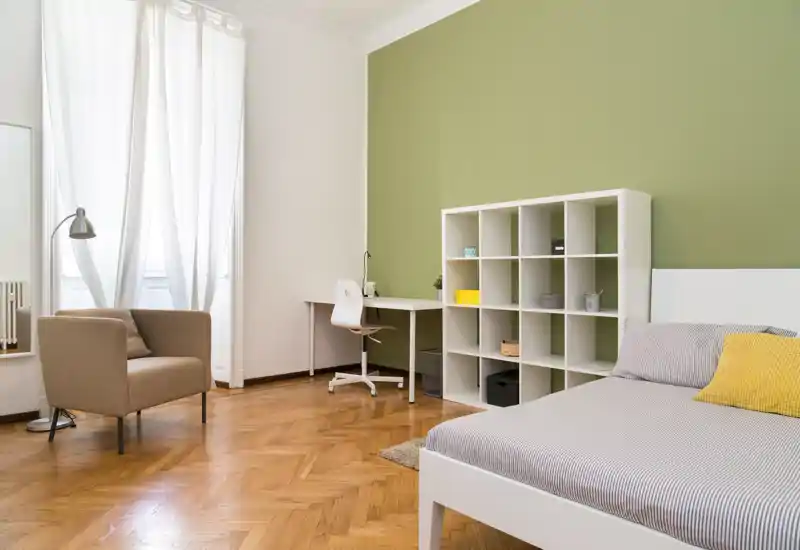 Bright, modern bedroom with a neatly made bed, open shelving unit and a desk by the window — minimalist, warm styling and plenty of natural light.