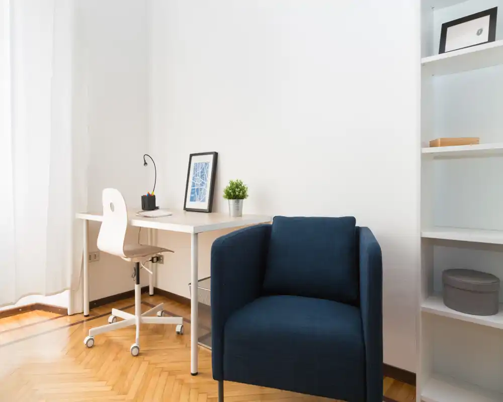 Zona giorno luminosa e minimal con una poltrona blu scuro, una scrivania semplice e luce naturale da una grande finestra — ordinata, moderna e confortevole per lavorare o rilassarsi.