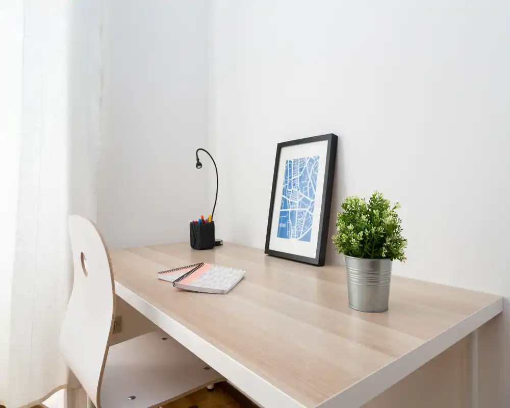 Angolo lavoro luminoso e minimale con scrivania in legno, sedia bianca, lampada da tavolo, stampa incorniciata e pianta in vaso — un tranquillo spazio studio domestico.