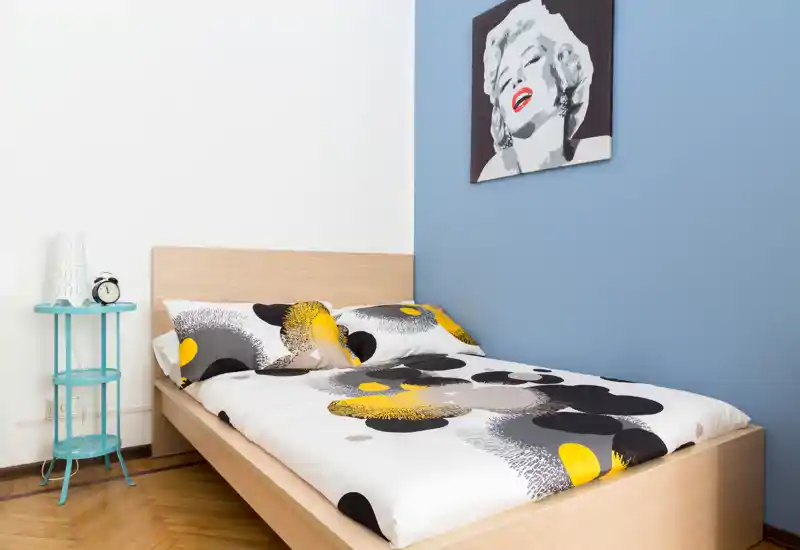 Camera da letto singola luminosa e moderna con struttura letto in legno, copriletto decorato e quadro a parete; comodino minimal e di design completa l’ambiente accogliente.