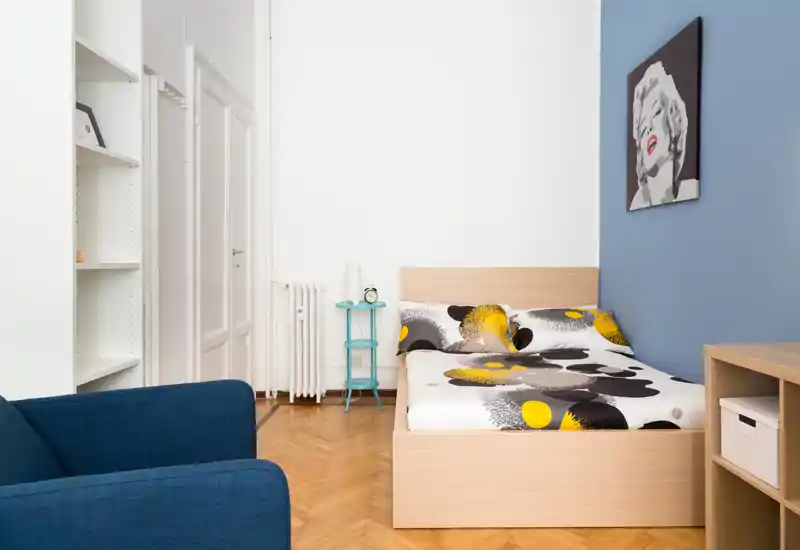 Camera da letto moderna e luminosa con letto matrimoniale in legno, biancheria stampata, parete azzurra e divano coordinato; ambiente ordinato e ben illuminato, perfetto per annunci.