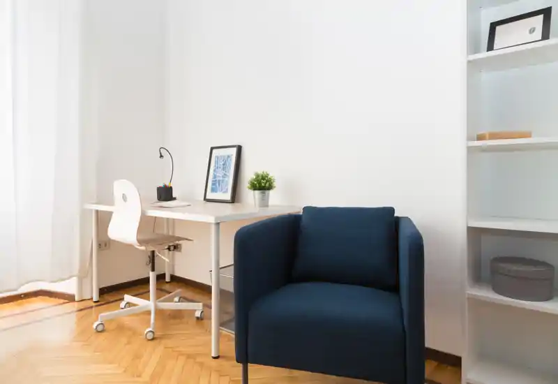 Zona giorno luminosa e minimal con una poltrona blu scuro, una scrivania semplice e luce naturale da una grande finestra — ordinata, moderna e confortevole per lavorare o rilassarsi.