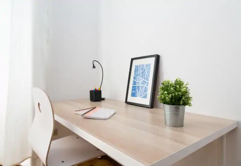 Angolo lavoro luminoso e minimale con scrivania in legno, sedia bianca, lampada da tavolo, stampa incorniciata e pianta in vaso — un tranquillo spazio studio domestico.