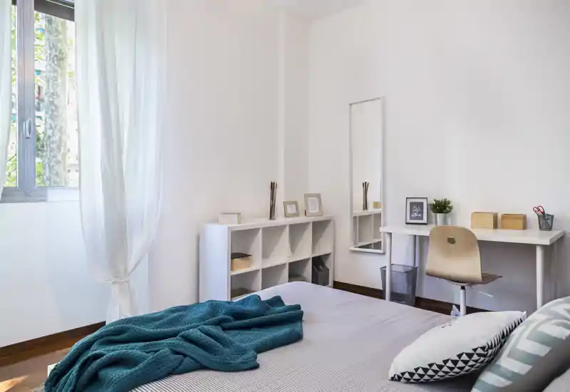 Chambre lumineuse et minimaliste avec lit bien fait, plaid bleu-vert et coussins décoratifs, étagères et petit bureau contre le mur — idéale pour un séjour calme et fonctionnel.
