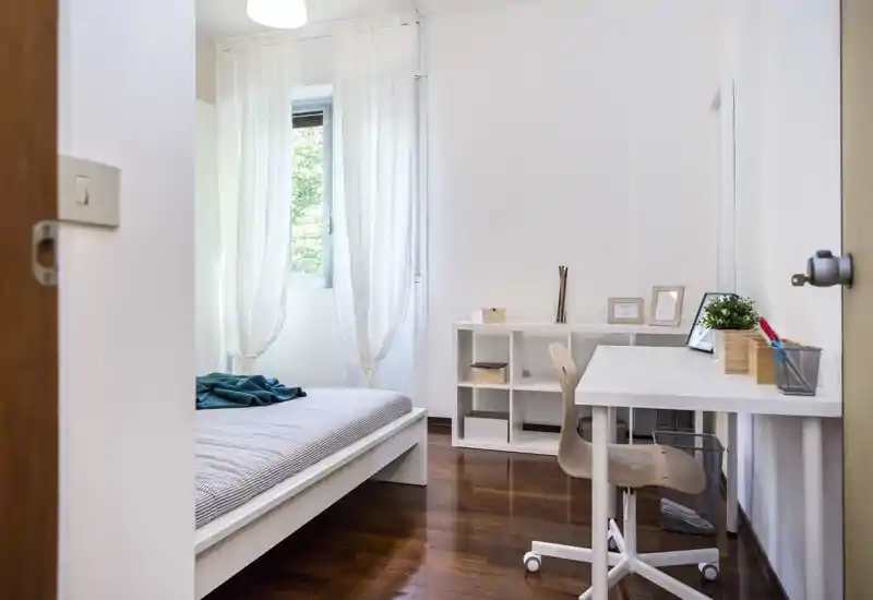Bright, minimal bedroom with a white bed frame, striped bedding and a functional desk next to shelving — calm, airy feel with natural light from the window.