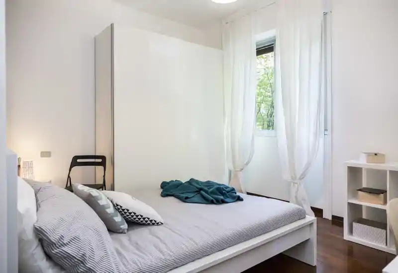 Chambre lumineuse et minimaliste avec un lit double bien fait, textiles doux et grande armoire. La lumière naturelle et la décoration blanche offrent une atmosphère calme et aérée.