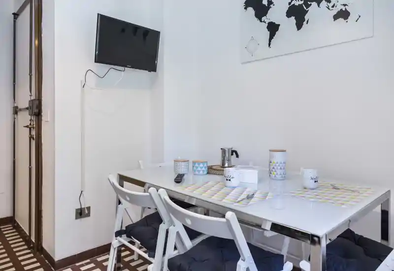 Bright, modern dining corner with a glass table set for casual dining, foldable chairs with cushions, and wall-mounted TV — ideal for a compact apartment.