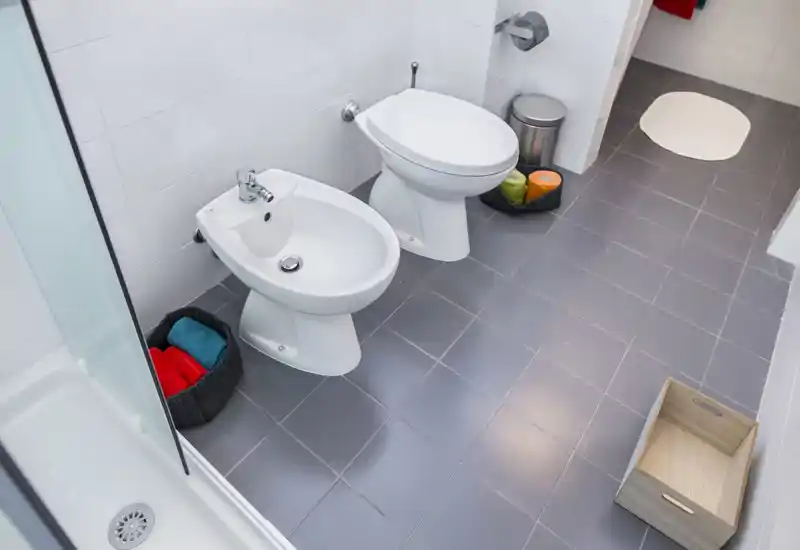 Clean, modern bathroom with a toilet and bidet, grey tiled floor, shower tray at the frame edge and neatly placed accessories.