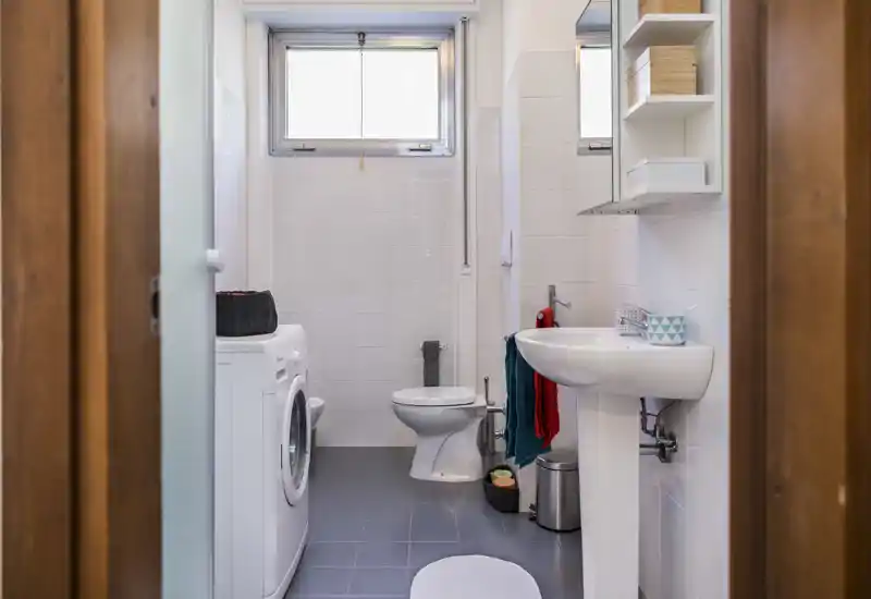 Bright, compact bathroom with a toilet, pedestal sink, mirror cabinet and a washing machine; tiled walls and floor provide a clean, functional space.