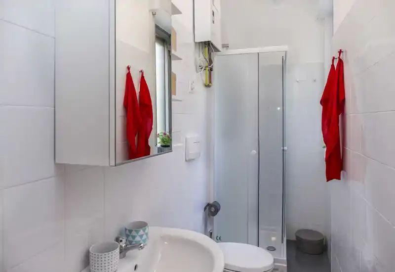 Bright, compact bathroom with a corner shower, wall-mounted sink and mirrored cabinet; clean white tiles and red towels add contrast.