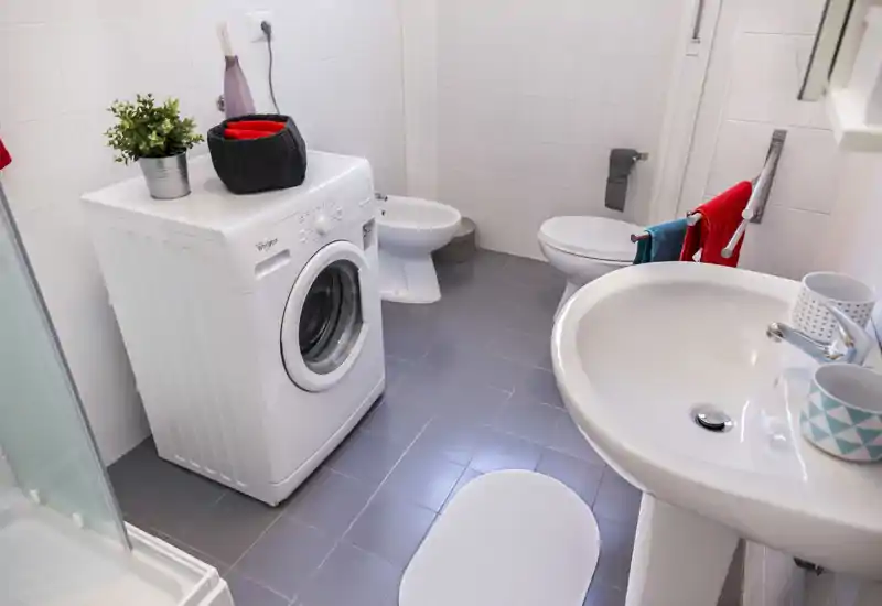 Bright, tidy bathroom with a sink, toilet, bidet and a washing machine neatly placed on the tiled floor.